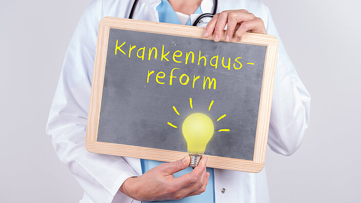 Eine ärztlich gekleidete Person hält eine Kreidetafel mit dem Wort "Krankenhausreform" darauf und eine Glühlampe hoch. 