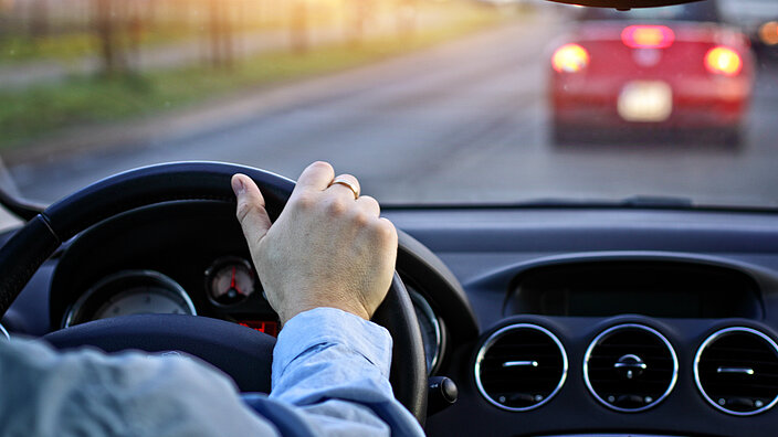 Blick aus der Frontscheibe eines Autos. Der Fahrer hält das Lenkrad. Vor ihm fährt ein rotes Auto. 