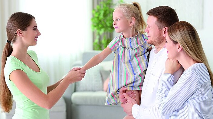 Eine Familie in einer Beratungssituation. Das Kind, ein Mädchen, ist auf dem Arm des Vaters. Gegenüber steht eine Familienberaterin, die die Hand des Mädchens hält.