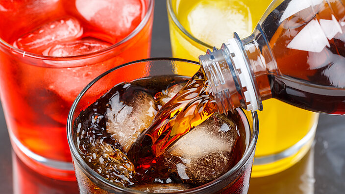 3 Gläser mit bunten Softdrinks. In ein Glas wird gerade noch Cola gegossen.