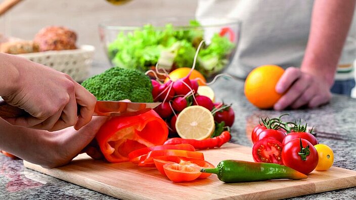 Auf einem Holzbrett sind verschiedene Gemüsesorten zu erkennen: Tomaten, Peperoni, Brokkoli, Radieschen. Eine rote Paprika wird in Scheiben geschnitten.