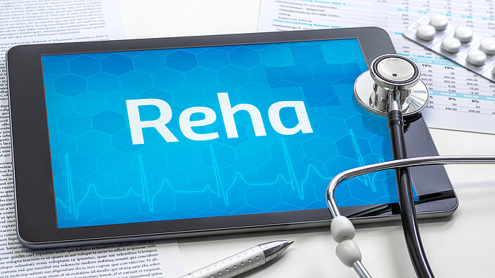 Auf einem Tablet steht das Wort "Reha". Im Hintergrund sind medizinische Befunde zu sehen, neben Tabletten, einem Stetoskop und einem Kugelschreiber.