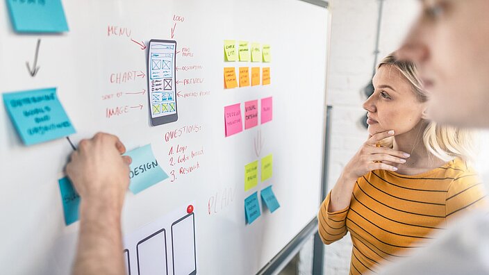 Ein Mann und eine Frau stehen vor einem Whiteboard an dem viele Notizen und Zettel hängen. Er zeigt auf einen Zettel und sie hat die Hand am Kinn.